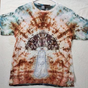 May include: A tie-dye t-shirt with a mushroom design in shades of brown, blue, and white. The shirt has a circular pattern with a mushroom in the center.