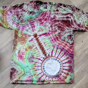 May include: A tie-dye t-shirt with a banjo design. The shirt features a vibrant mix of colors, including shades of green, purple, brown, and white. The banjo is centrally placed, with a brown neck and a circular body. The shirt has short sleeves and a crew neck.