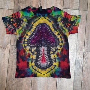 May include: A tie-dye t-shirt featuring a large mushroom graphic. The mushroom is purple with white dots and a red stem. The shirt has a colorful, psychedelic design with shades of red, yellow, blue, and green.