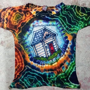 May include: Tie-dye t-shirt with a colorful spiral design in orange, green, and blue. The shirt features a detailed illustration of a small house with a red door and blue roof, surrounded by green foliage.