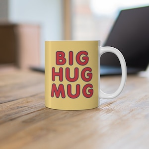 May include: A white ceramic mug with a yellow background and the text "BIG HUG MUG" in red letters with a blue outline.