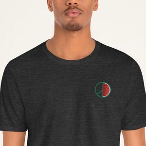 Può includere: T-shirt grigio scuro con un piccolo disegno ricamato di un simbolo di pace con una fetta di anguria all'interno. L'anguria è rossa e verde. La maglietta è a girocollo.
