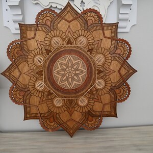 May include: A large, intricately carved wooden mandala wall art piece. The design features a central starburst pattern surrounded by floral and geometric elements. The wood has varying shades of brown, creating depth and visual interest. The mandala is displayed on a light-colored wall.
