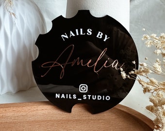 May include: Black and white round sign with the text "Nails by Amelia" and "Nails_Studio" with an Instagram icon. The sign has a scalloped edge.