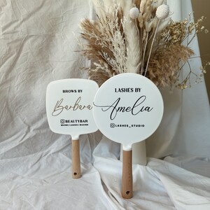 May include: Two white handheld mirrors with wooden handles. The top mirror is round and says "Lashes by Amelia" with a social media icon and the text "Lashes_Studio". The bottom mirror is square and says "Brows by Barbara" with a social media icon and the text "Beautybar Brows | Lashes | Waxing".
