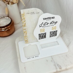 Peut inclure: Un pr&eacute;sentoir en acrylique blanc avec une inscription dor&eacute;e qui dit "Let's stay connected". Le pr&eacute;sentoir comporte deux codes QR et une &eacute;tiquette "Tap to Pay". Le pr&eacute;sentoir comporte &eacute;galement une inscription dor&eacute;e qui dit "These girls sold out".