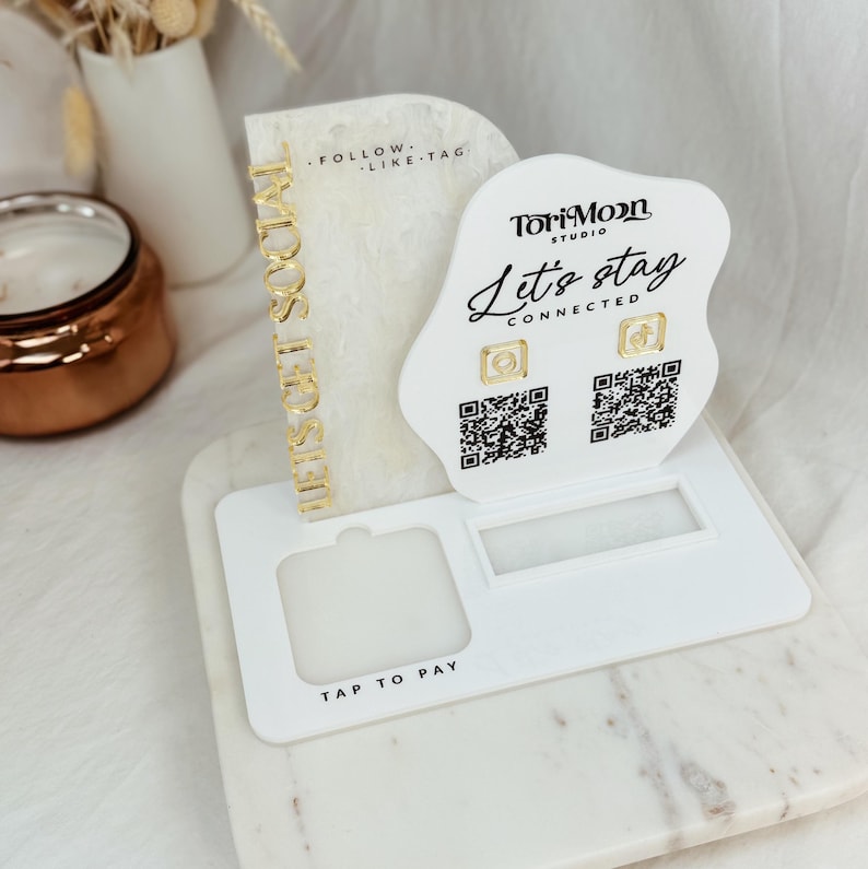 Peut inclure: Un pr&eacute;sentoir en acrylique blanc avec une inscription dor&eacute;e qui dit "Let's stay connected". Le pr&eacute;sentoir comporte deux codes QR et une &eacute;tiquette "Tap to Pay". Le pr&eacute;sentoir comporte &eacute;galement une inscription dor&eacute;e qui dit "These girls sold out".