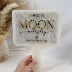 May include: A white and cream-colored rectangular sign with a handle, featuring the text "Lashes By Moon Artistry" in gold and black. The sign has a marbled effect and is held by a hand.