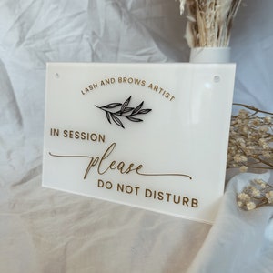 May include: White acrylic sign with gold lettering that reads "Lash and Brows Artist In Session Please Do Not Disturb". A small black twig with leaves is above the text "In Session".