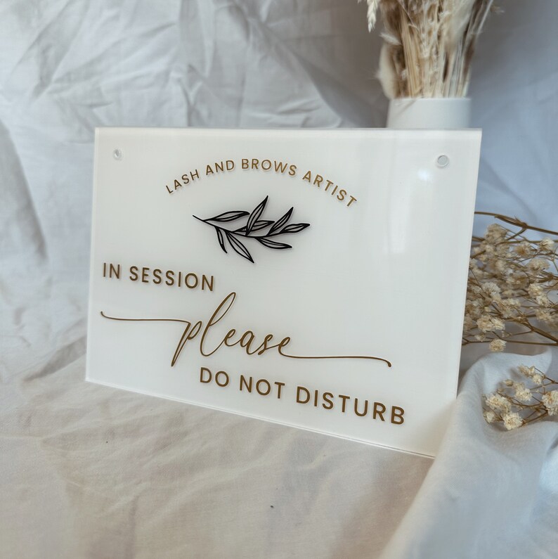 May include: White acrylic sign with gold lettering that reads "Lash and Brows Artist In Session Please Do Not Disturb". A small black twig with leaves is above the text "In Session".