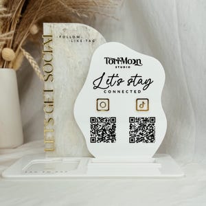 Peut inclure: Un pr&eacute;sentoir blanc avec une pancarte blanche et dor&eacute;e qui dit "Let's Get Social" et "ToriMoon Studio" avec le texte "Let's stay connected" et des ic&ocirc;nes pour Instagram et TikTok. Des codes QR sont &eacute;galement inclus pour chaque plateforme de m&eacute;dias sociaux.