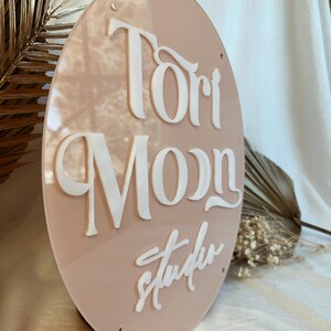May include: A round, light pink acrylic sign with the text "Tori Moon Studio" in white raised lettering.