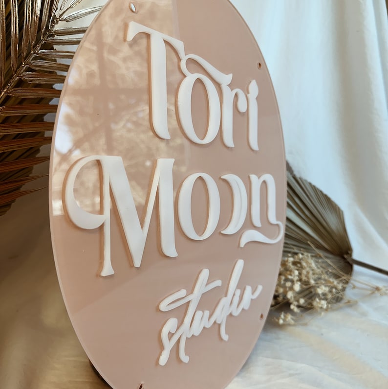 May include: A round, light pink acrylic sign with the text "Tori Moon Studio" in white raised lettering.
