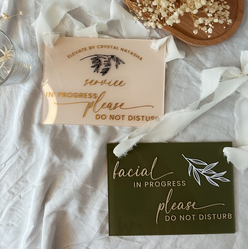 May include: Two acrylic signs with gold lettering and a leafy design. The top sign is pink and says "service in progress please do not disturb". The bottom sign is green and says "facial in progress please do not disturb".