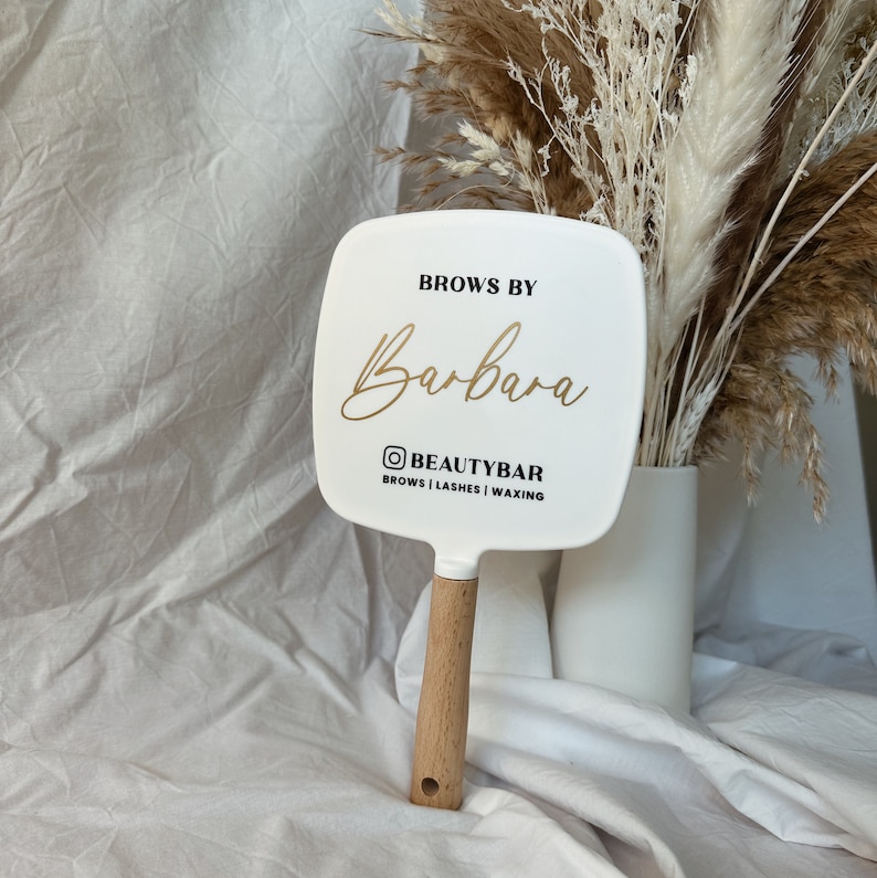 Peut inclure: Un miroir &agrave; main blanc avec un manche en bois. Le miroir porte l'inscription "Brows by Barbara" en lettres dor&eacute;es, ainsi que le compte Instagram "@BEAUTYBAR" et les services propos&eacute;s : sourcils, cils et &eacute;pilation.