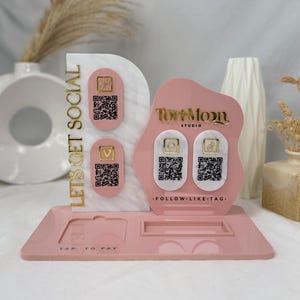 May include: A pink acrylic social media display stand with gold lettering and QR codes. The stand features the text "LET'S GET SOCIAL" and "TAP TO PAY". The stand has a slot for a card and two areas for QR codes.