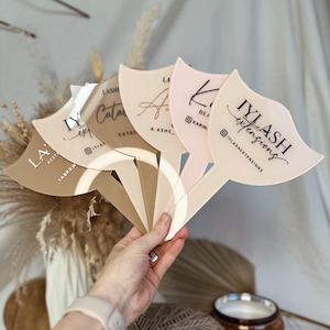 May include: Five acrylic signs with different business names and social media handles. The signs are shaped like a fan and are in various shades of brown and pink.
