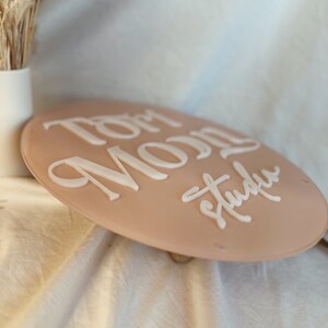 May include: A round, light pink sign with the words "Tom Moon Studio" written in white raised lettering.