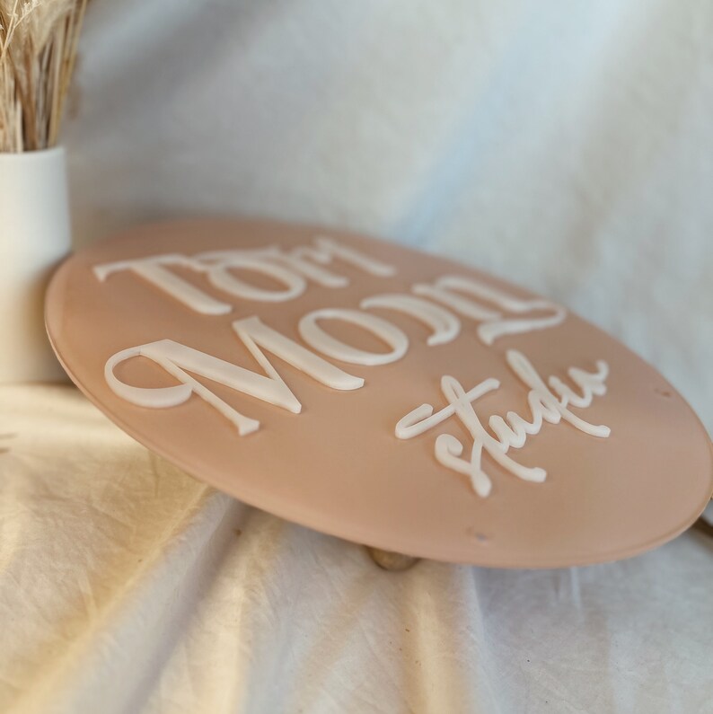 May include: A round, light pink sign with the words "Tom Moon Studio" written in white raised lettering.