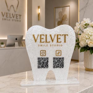 May include: A white marble-patterned acrylic sign shaped like a tooth, featuring the gold text VELVET SMILE STUDIO, an Instagram icon, a TikTok icon, and two black QR codes. The sign stands on a rectangular base of the same material.