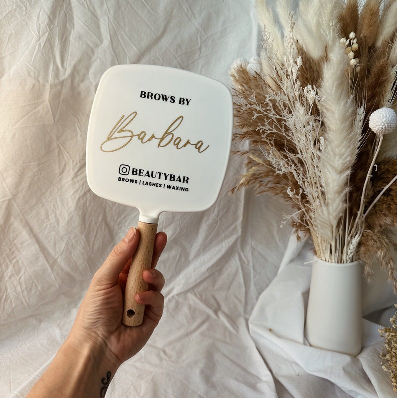Peut inclure: Miroir &agrave; main blanc avec manche en bois. Le miroir porte l'inscription "Brows by Barbara" en lettres dor&eacute;es, ainsi que le nom d'utilisateur Instagram "@BEAUTYBAR" et les services propos&eacute;s. Le miroir est pr&eacute;sent&eacute; sur fond de fleurs s&eacute;ch&eacute;es.