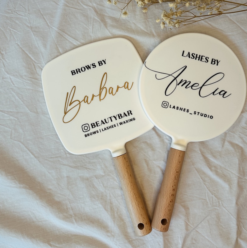 Peut inclure: Deux miroirs &agrave; main blancs avec des manches en bois. L'un est carr&eacute; avec "Brows by Barbara" en &eacute;criture dor&eacute;e et le texte "BEAUTYBAR". L'autre est rond avec "Lashes by Amelia" en &eacute;criture noire et le texte "LASHES_STUDIO".