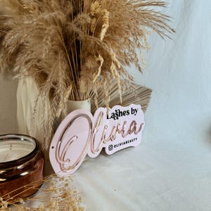 May include: Pink acrylic sign with the text "Lashes by Olivia" in rose gold lettering and the Instagram handle "@oliviabeauty" below. The sign is propped up against a vase of dried pampas grass.
