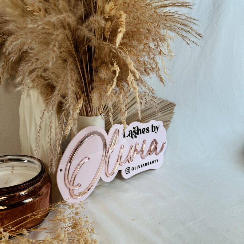May include: Pink acrylic sign with the text "Lashes by Olivia" in rose gold lettering and the Instagram handle "@oliviabeauty" below. The sign is propped up against a vase of dried pampas grass.