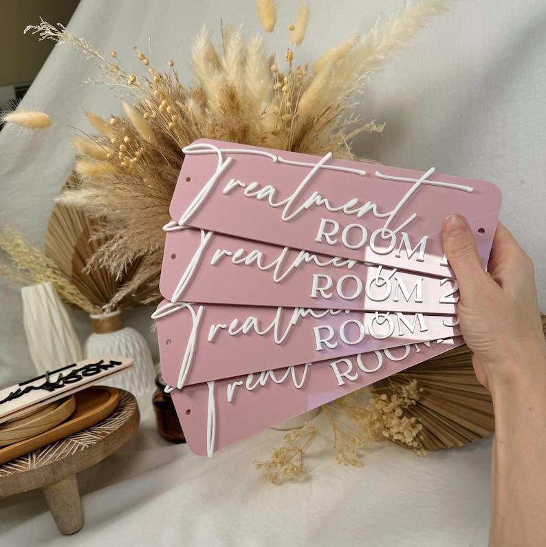 Op de afbeelding: Vijf roze acrylplaten met witte letters die "Behandelkamer 1", "Behandelkamer 2", "Behandelkamer 3", "Behandelkamer 4" en "Behandelkamer 5" zeggen.