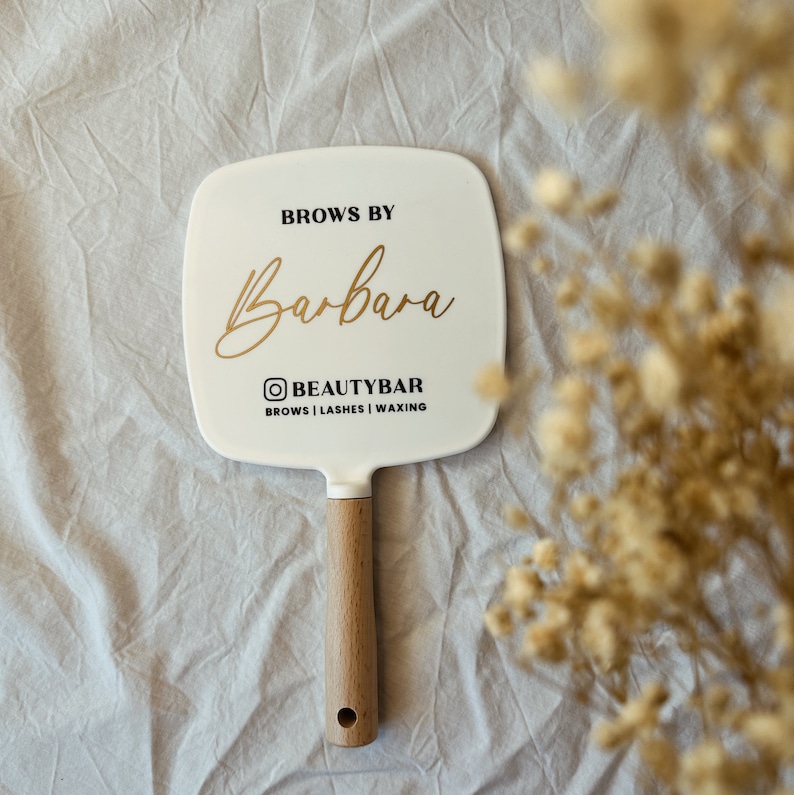 Peut inclure: Un miroir &agrave; main blanc avec un manche en bois. Le miroir porte l'inscription "Brows by Barbara" en lettres dor&eacute;es, avec le nom d'utilisateur Instagram "@BEAUTYBAR" et les services propos&eacute;s : "Brows | Lashes | Waxing" en noir.
