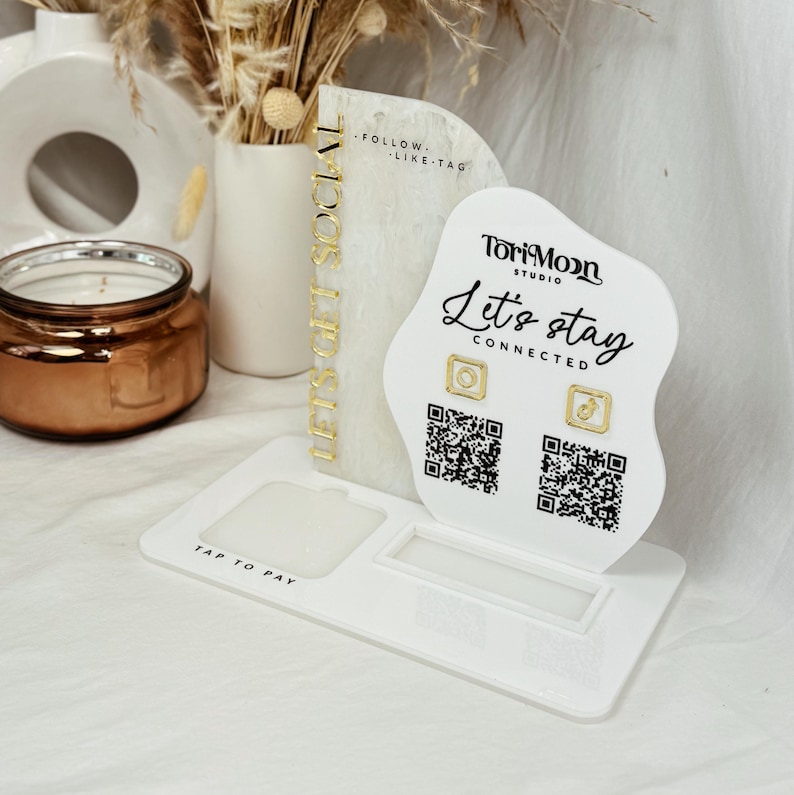 Peut inclure: Un pr&eacute;sentoir en acrylique blanc avec une inscription dor&eacute;e qui dit "Let's Get Social". Le pr&eacute;sentoir a une d&eacute;coupe pour un t&eacute;l&eacute;phone et une section avec le texte "TorriMoon Studio" et "Let's stay connected". Il y a deux codes QR et deux ic&ocirc;nes dor&eacute;es.