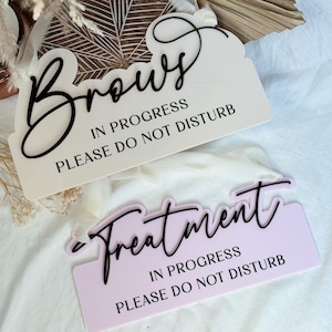 May include: Two signs with the text "Brows in progress, please do not disturb" and "Treatment in progress, please do not disturb". The top sign is white with black text and the bottom sign is light purple with black text.