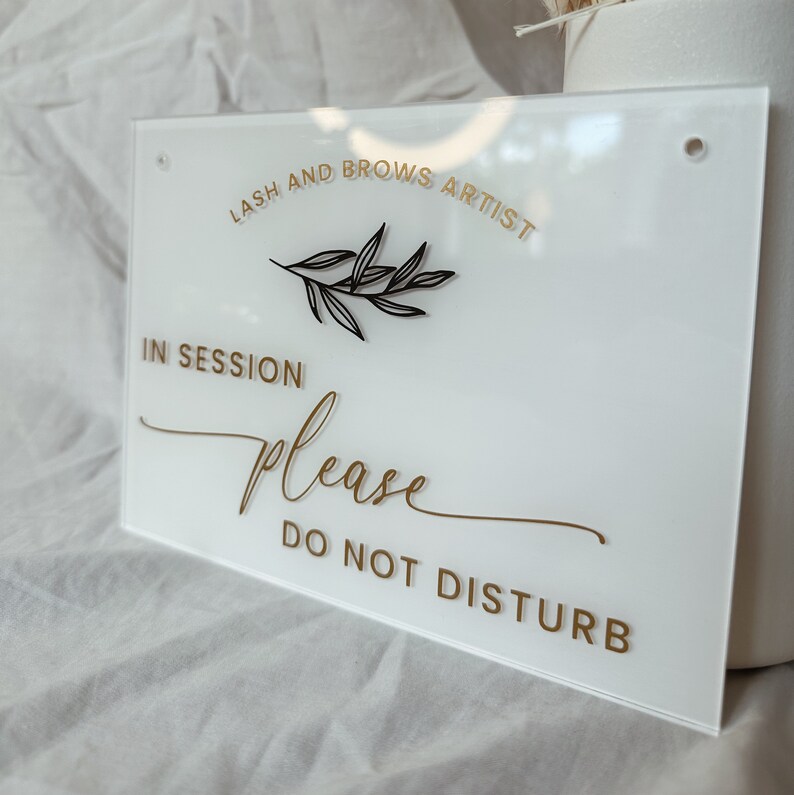 May include: White acrylic sign with gold lettering that reads "Lash and Brows Artist" with a small black branch design. The sign also reads "In Session Please Do Not Disturb".