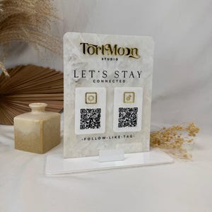 May include: A white and gold acrylic display stand with the text "Tori Moon Studio" and "Let's Stay Connected." It features two QR codes with social media icons and the phrase "Follow. Like. Tag."