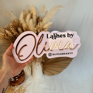 May include: Pink acrylic sign with the text "Lashes by Olivia" in gold lettering. The sign also includes the Instagram handle "@oliviabeauty".