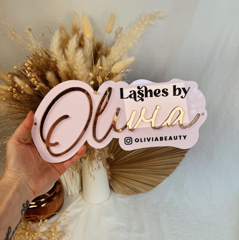 May include: Pink acrylic sign with the text "Lashes by Olivia" in gold lettering. The sign also includes the Instagram handle "@oliviabeauty".