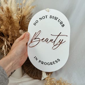May include: White round sign with black text that reads "Do Not Disturb Beauty In Progress". The word "Beauty" is written in cursive in rose gold.