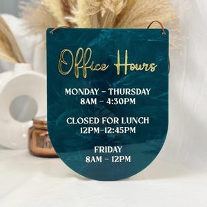May include: A teal sign with gold lettering that reads "Office Hours." The sign displays the business hours: Monday-Thursday 8 AM-4:30 PM, closed for lunch 12 PM-12:45 PM, and Friday 8 AM-12 PM. The sign has a leather cord for hanging.