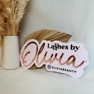 May include: Pink and white sign with the text "Lashes by Olivia" in black lettering and a rose gold cursive Olivia. The sign also includes the Instagram handle "@OliviaBeauty".