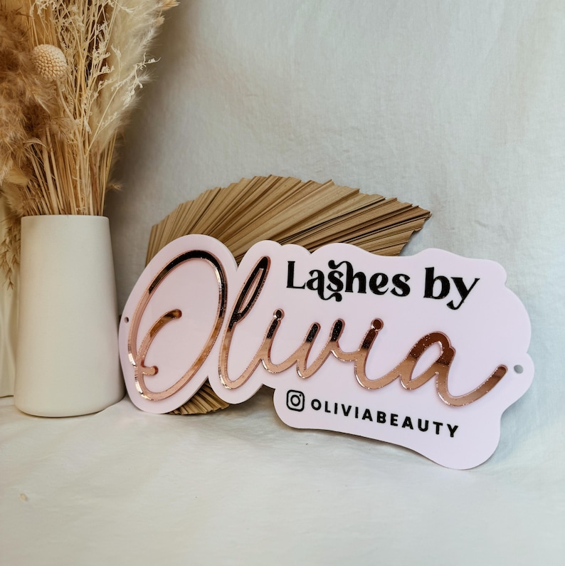 May include: Pink and white sign with the text "Lashes by Olivia" in black lettering and a rose gold cursive Olivia. The sign also includes the Instagram handle "@OliviaBeauty".