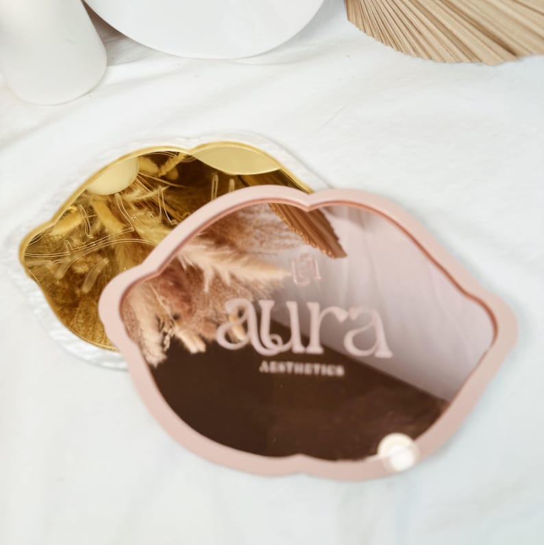 May include: Two pink and gold lip-shaped decorative mirrors. The larger mirror has the text "aura aesthetics" printed on it.