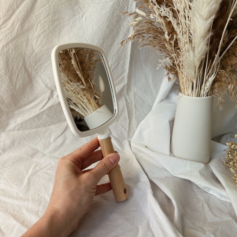 Peut inclure: Un miroir &agrave; main rectangulaire blanc cass&eacute; avec un manche en bois. Le miroir refl&egrave;te un vase de fleurs s&eacute;ch&eacute;es. La poign&eacute;e est en bois clair avec un petit trou &agrave; la base. Le miroir a des coins arrondis et un cadre blanc.
