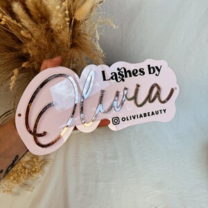 May include: Pink acrylic sign with the text "Lashes by Olivia" in black lettering and a rose gold glitter finish. The sign also includes the Instagram handle "@oliviabeauty".