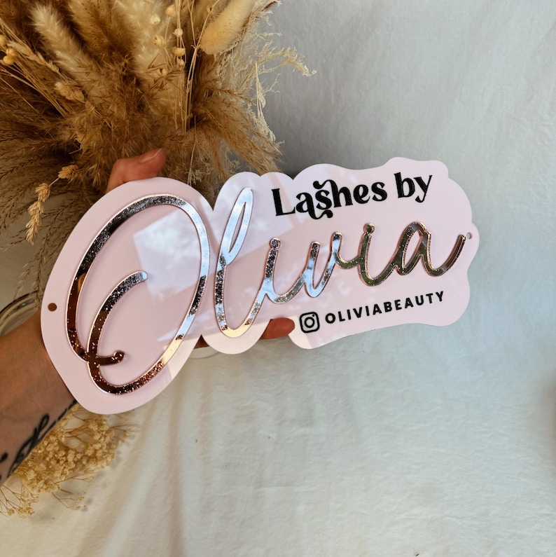 May include: Pink acrylic sign with the text "Lashes by Olivia" in black lettering and a rose gold glitter finish. The sign also includes the Instagram handle "@oliviabeauty".