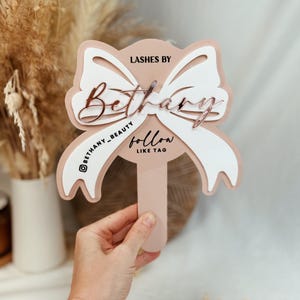 May include: A pink and white bow-shaped sign with the text "LASHES BY Bethany" and "@Bethany_Beauty follow like tag" in black and gold lettering.
