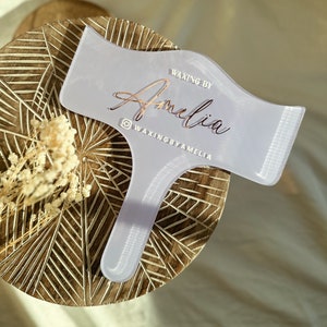 May include: A white plastic waxing tool with a handle and a curved edge. The tool has the text "WAXING BY Amelia" and "@WAXINGBYAMELIA" printed on it in rose gold.