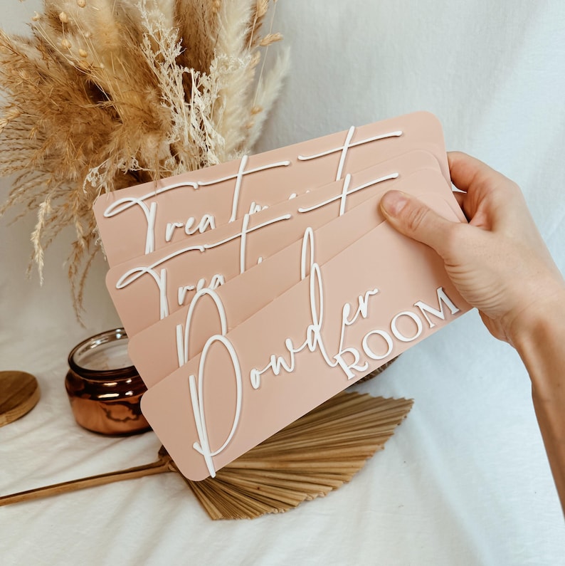 Op de afbeelding: Een set van vier roze houten borden met witte letters. De borden lezen "Treat Yourself", "Powder Room" en "Treat Yourself".
