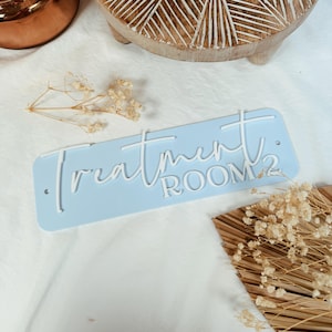 Op de afbeelding: Een lichtblauw rechthoekig bord met witte letters met de tekst "Treatment Room 2".