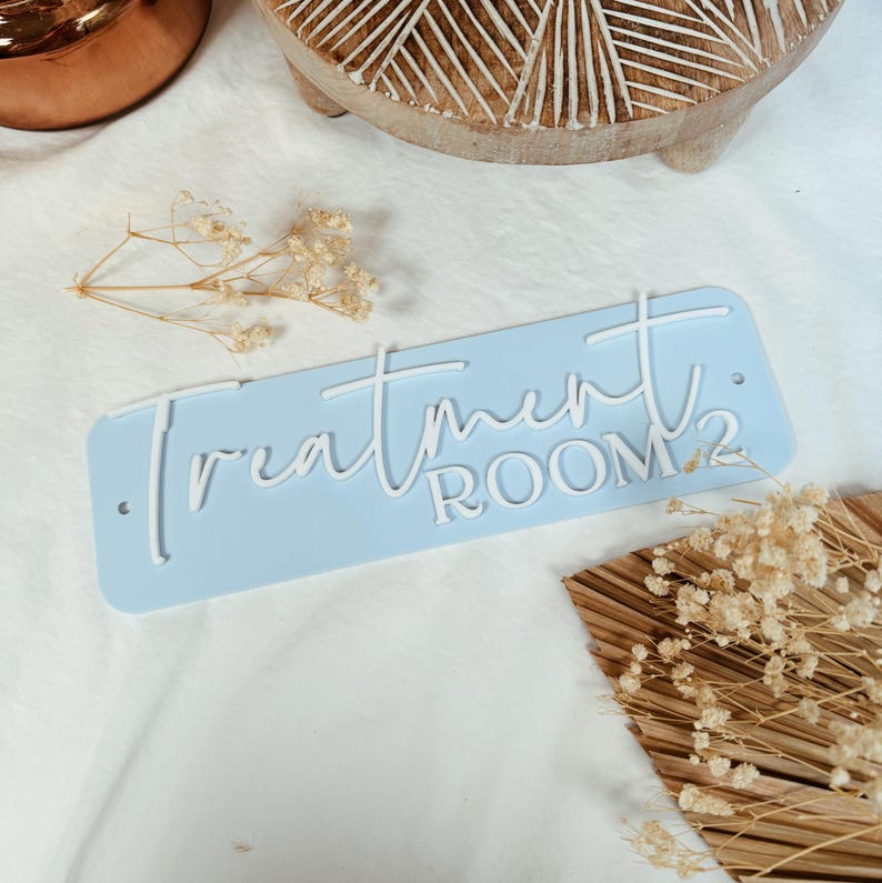 Op de afbeelding: Een lichtblauw rechthoekig bord met witte letters met de tekst "Treatment Room 2".