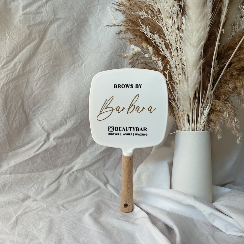 Peut inclure: Miroir &agrave; main blanc avec un manche en bois. Le miroir porte l'inscription "Brows by Barbara" en lettres dor&eacute;es, ainsi que le logo et les services de Beauty Bar. Le miroir est carr&eacute; avec des bords arrondis.
